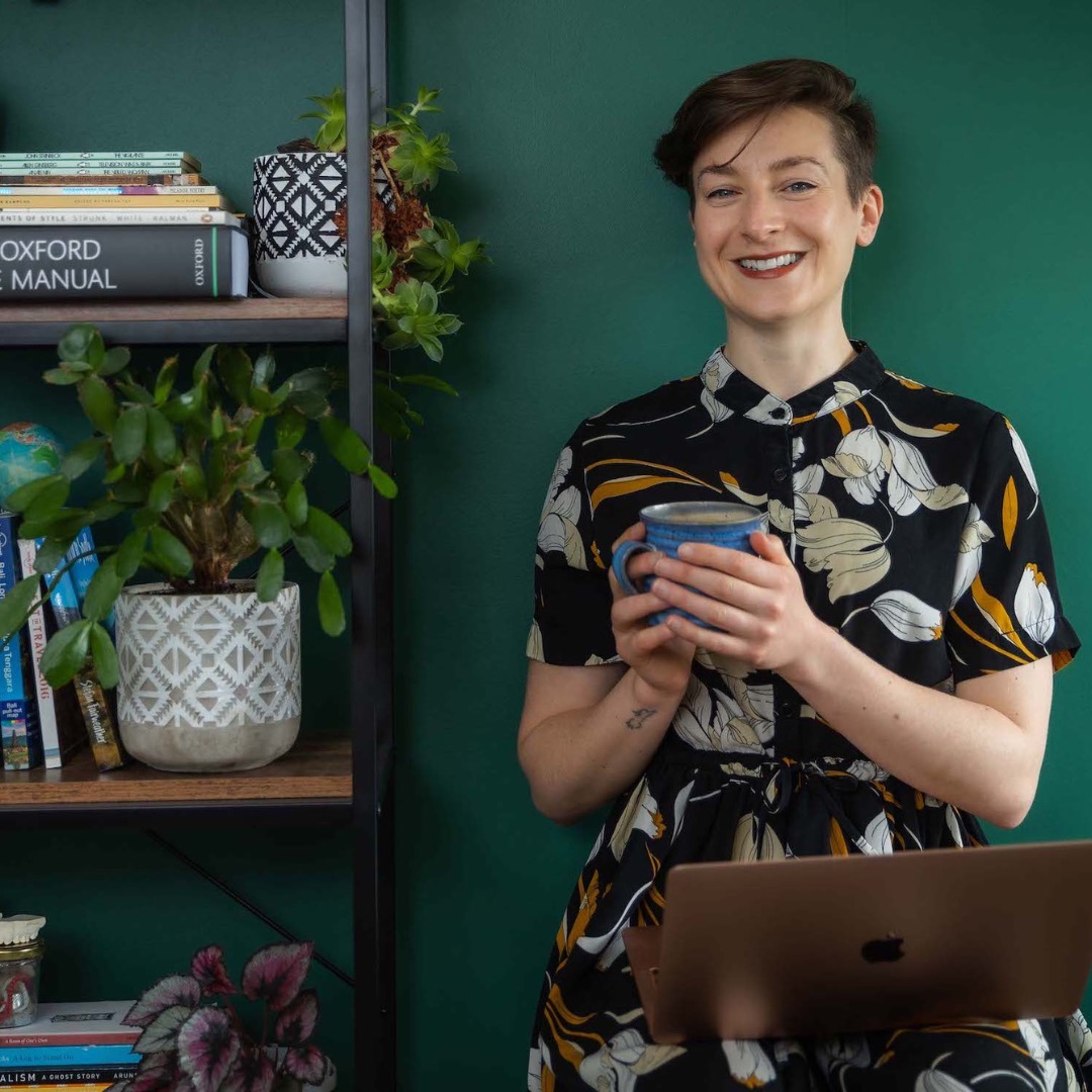 B G Rogers, author holds a cup of coffee next to a bookshelf in a room with a green wall